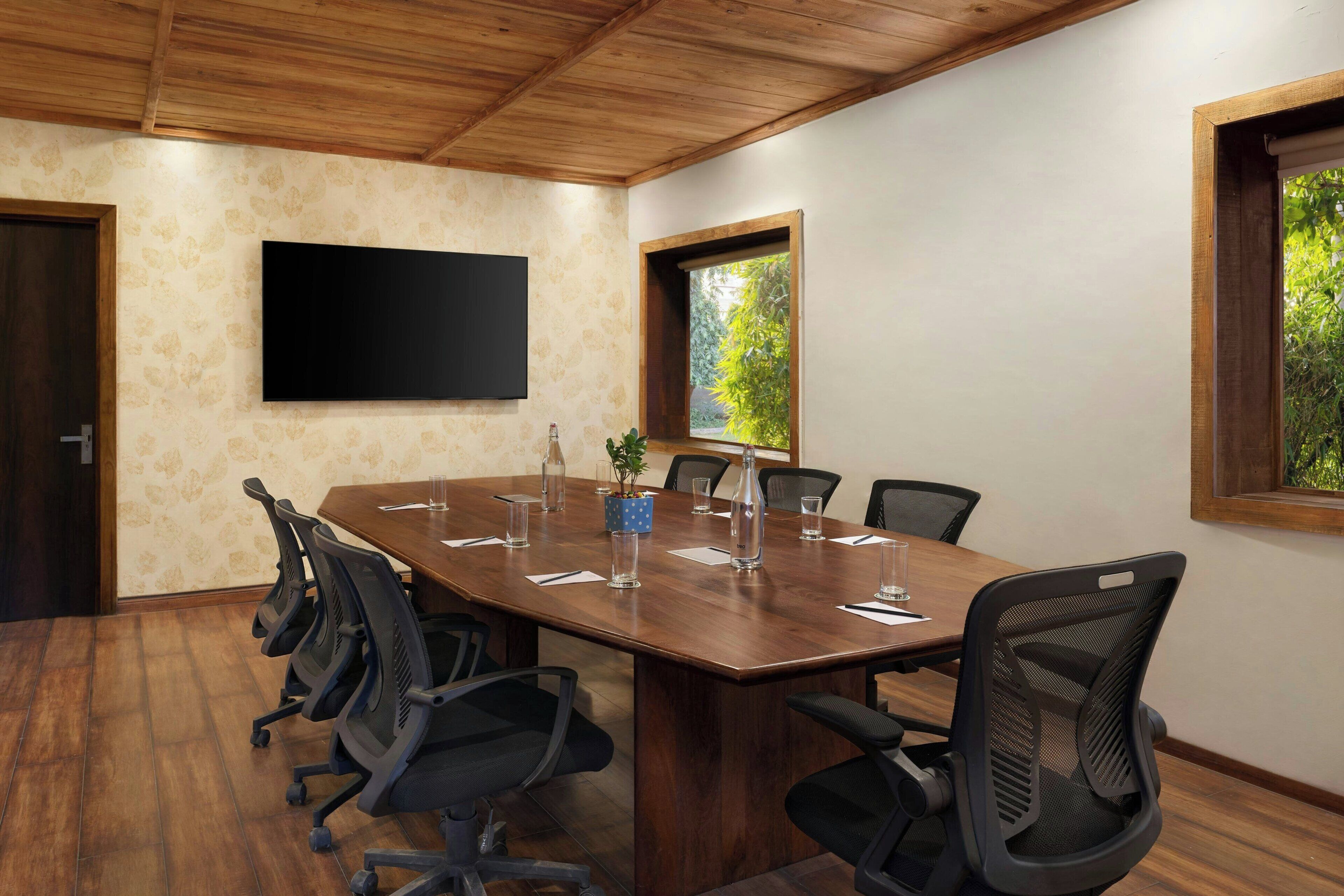 A conference room with a long table, chairs, a flat-screen TV, windows with views, and a wooden ceiling.