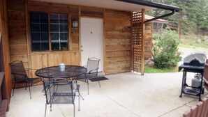 A wooden cabin with a patio featuring a round table and chairs, a grill, and a door with a number.
