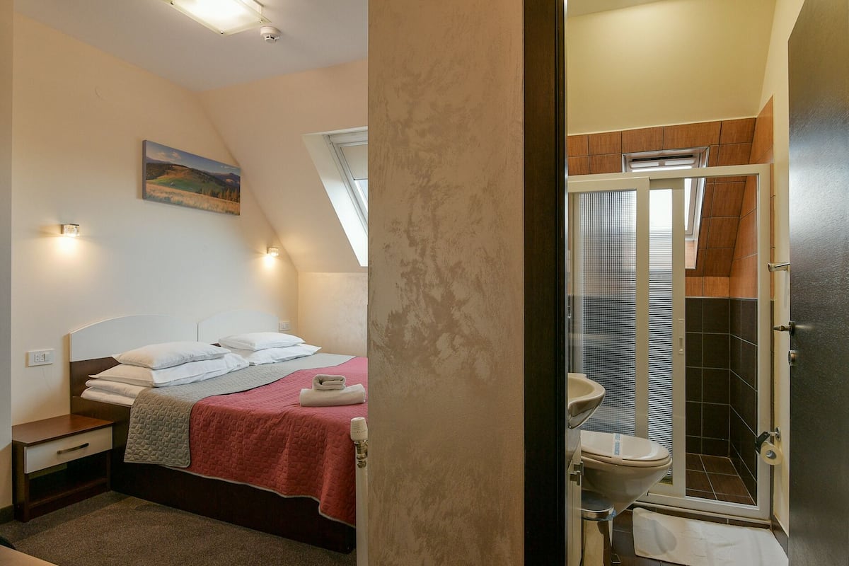 A bedroom with a bed, a nightstand, a painting on the wall, and a bathroom with a toilet, shower, and tiled walls.