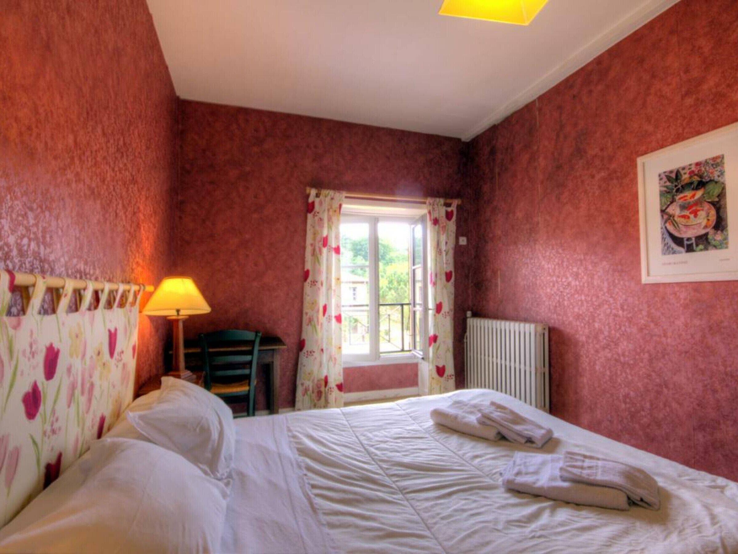A bedroom with a bed, a chair, a window with floral curtains, a radiator, and a lamp.
