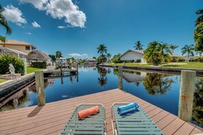 Une terrasse en bois avec deux chaises longues, dont une avec une serviette, donnant sur une étendue d’eau calme où se reflètent les palmiers et les maisons.