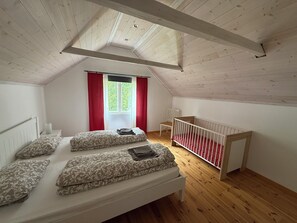 Ein Zimmer mit schrägen Wänden, einem Bett mit gemusterten Textilien, einem Babybett mit roter Bettwäsche und einem Fenster mit roten Vorhängen.