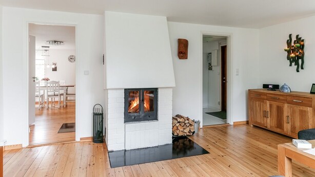 Een woonkamer met een witte open haard, houten vloeren, een eethoek op de achtergrond en een houten kast met een radio.