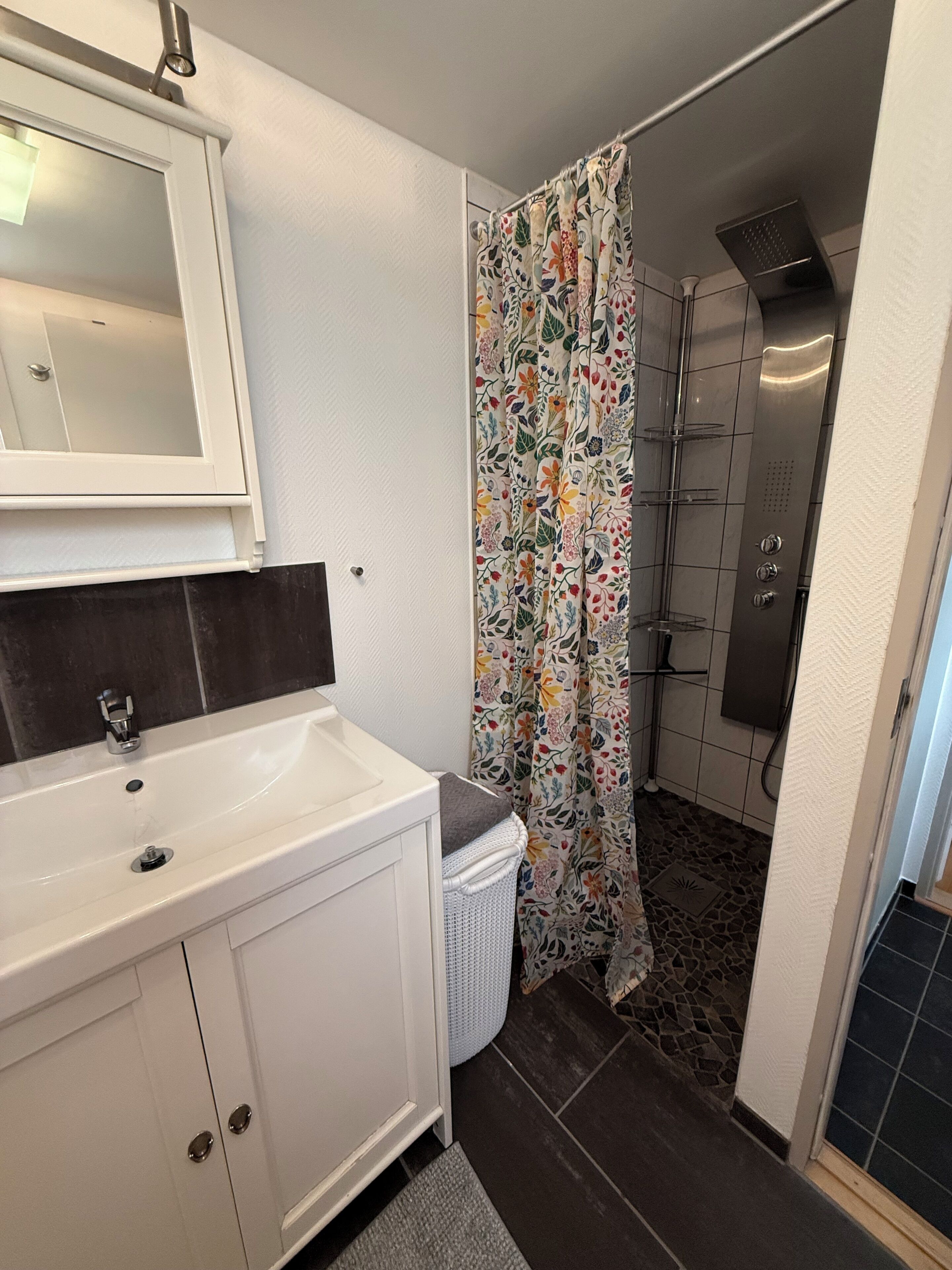 Une salle de bain avec un lavabo blanc, un miroir, une douche avec un rideau à motifs floraux et un sol carrelé.