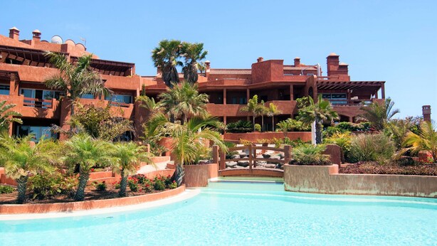 A resort with a swimming pool, palm trees, and a multi-story building with balconies.