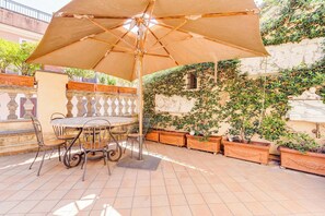 An outdoor patio with a table and chairs under a large umbrella, surrounded by potted plants and a stone wall with greenery. - Colosseum Terrace Apartment (Roma)
