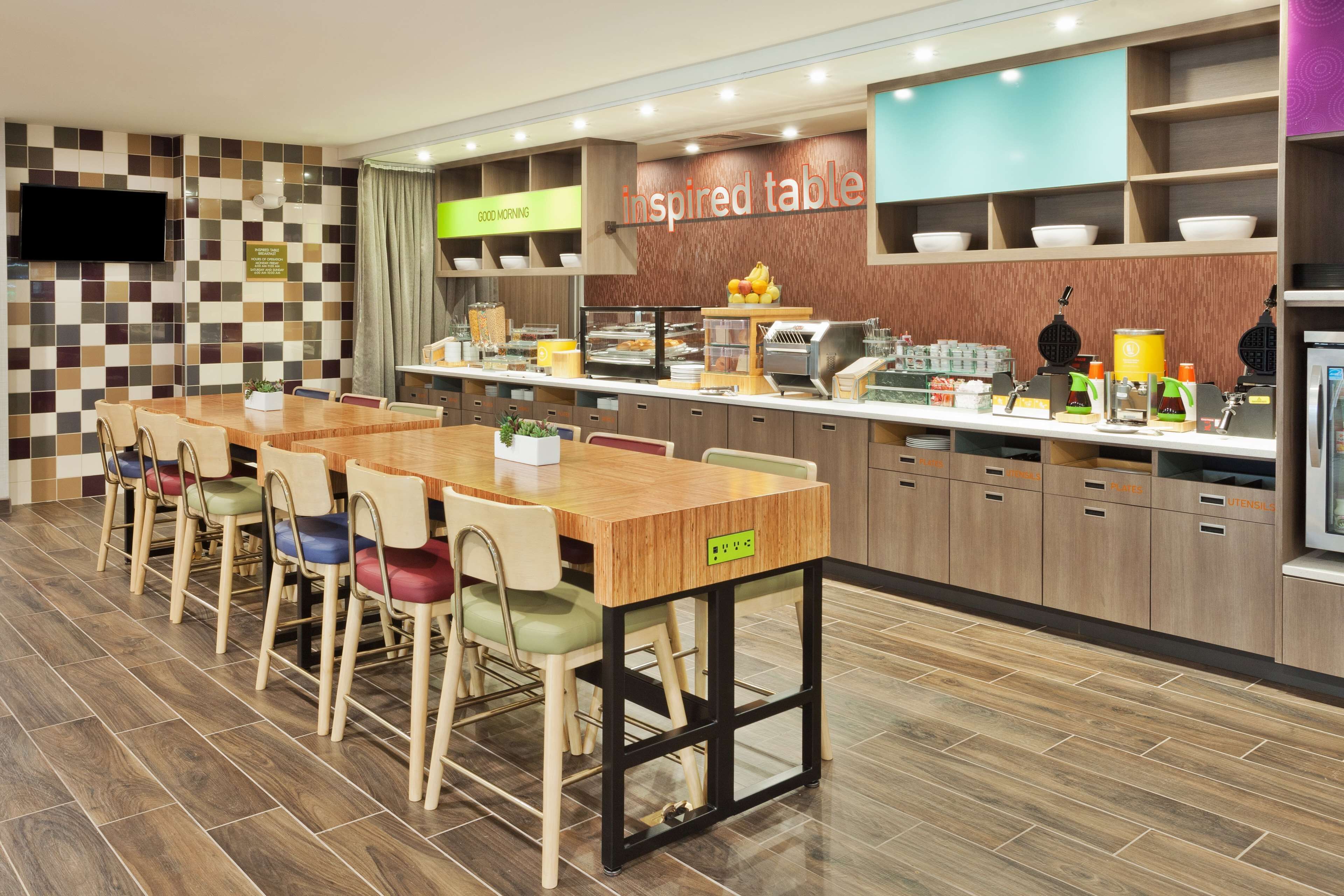 A modern restaurant with a checkered tile wall, a wooden table with chairs, and a counter with food items.
