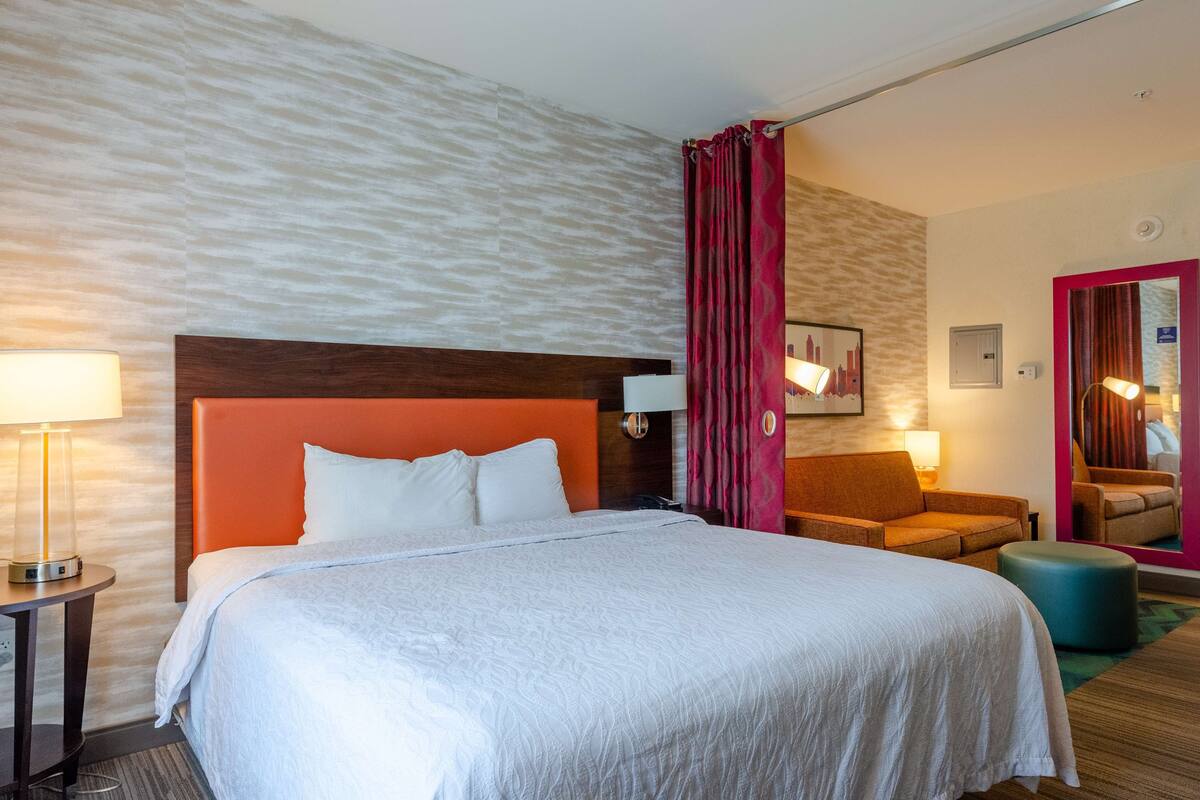 Habitación de hotel con una cama grande, cabecera de madera, mesita de noche con lámpara, zona de estar con una silla y reposapiés, y un espejo con marco rojo.