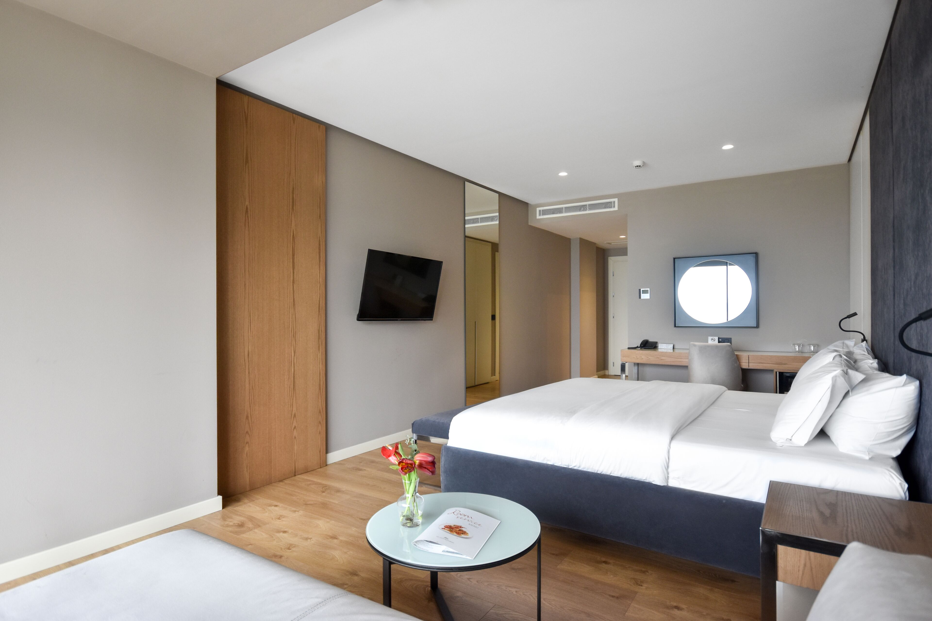 A modern hotel room with a large bed, a small round table with a vase, a desk with a chair, and a mirror on the wall.