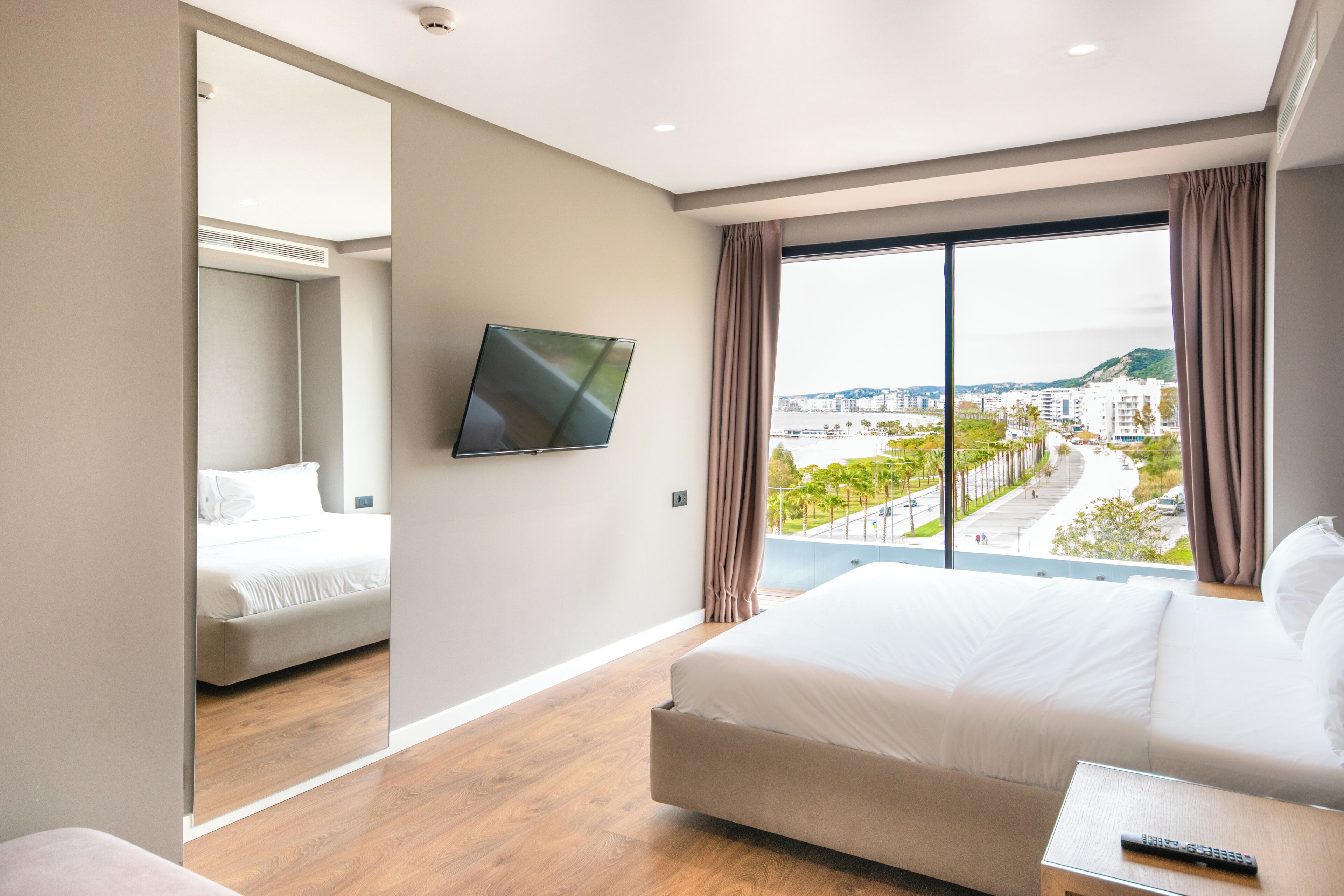 Een moderne hotelkamer met een groot bed, een aan de muur bevestigde televisie en uitzicht op de stad door grote ramen.