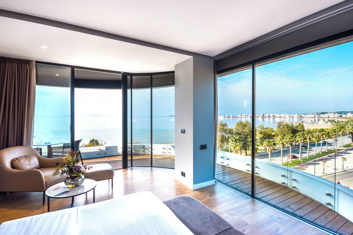A modern hotel room with a large balcony, a bed, a sofa, and a coffee table.