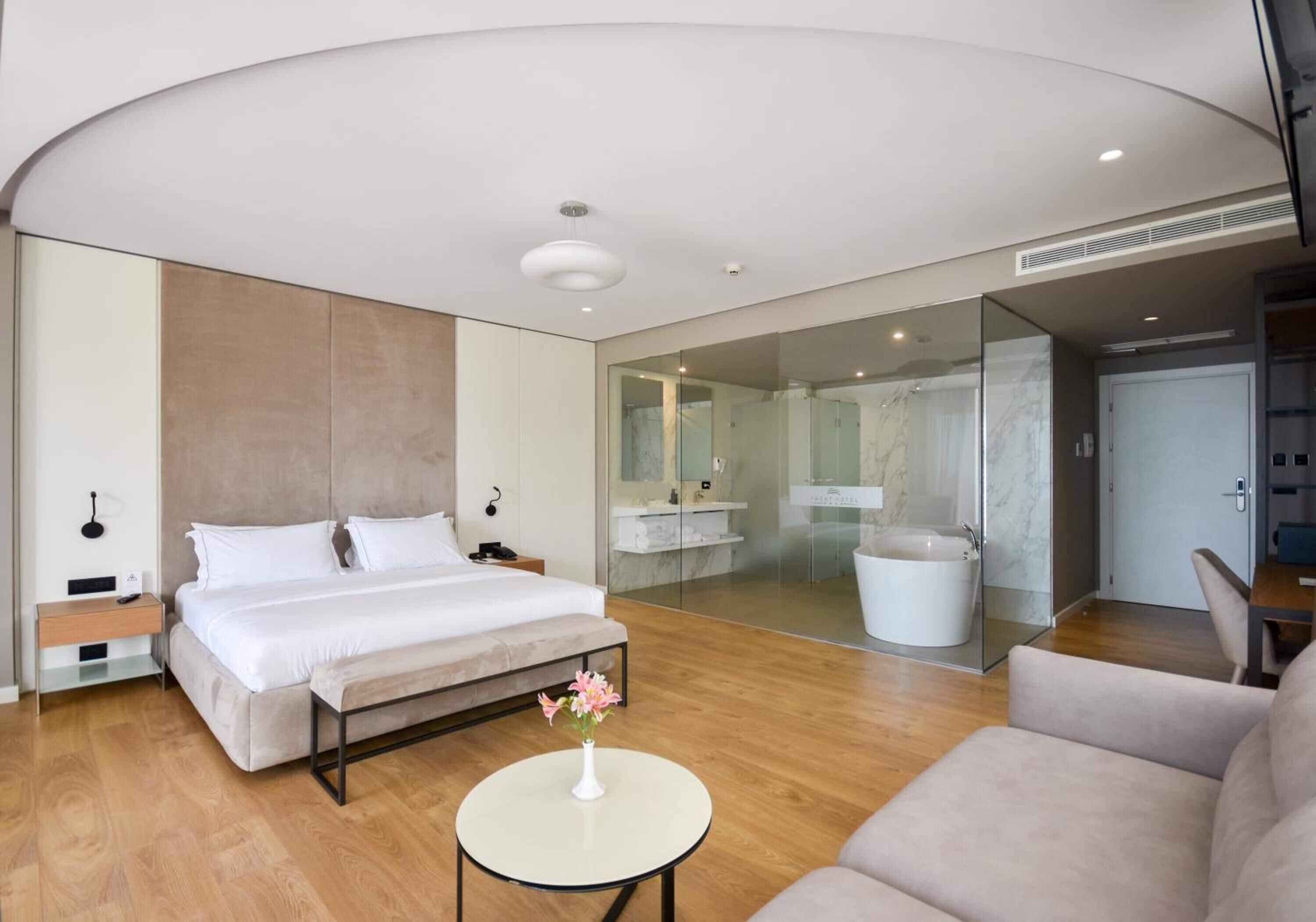 A modern hotel room with a large bed, a round coffee table with flowers, a sofa, a bathroom with a bathtub, and a glass shower enclosure.