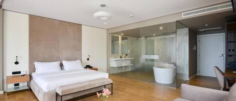 A modern hotel room with a large bed, a round coffee table with flowers, a sofa, a bathroom with a bathtub, and a glass shower enclosure.