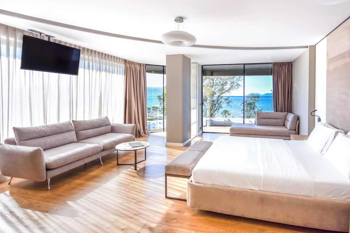 A modern hotel room with a large bed, a sofa, a coffee table, and a TV. The room has large windows with a view of the sea.