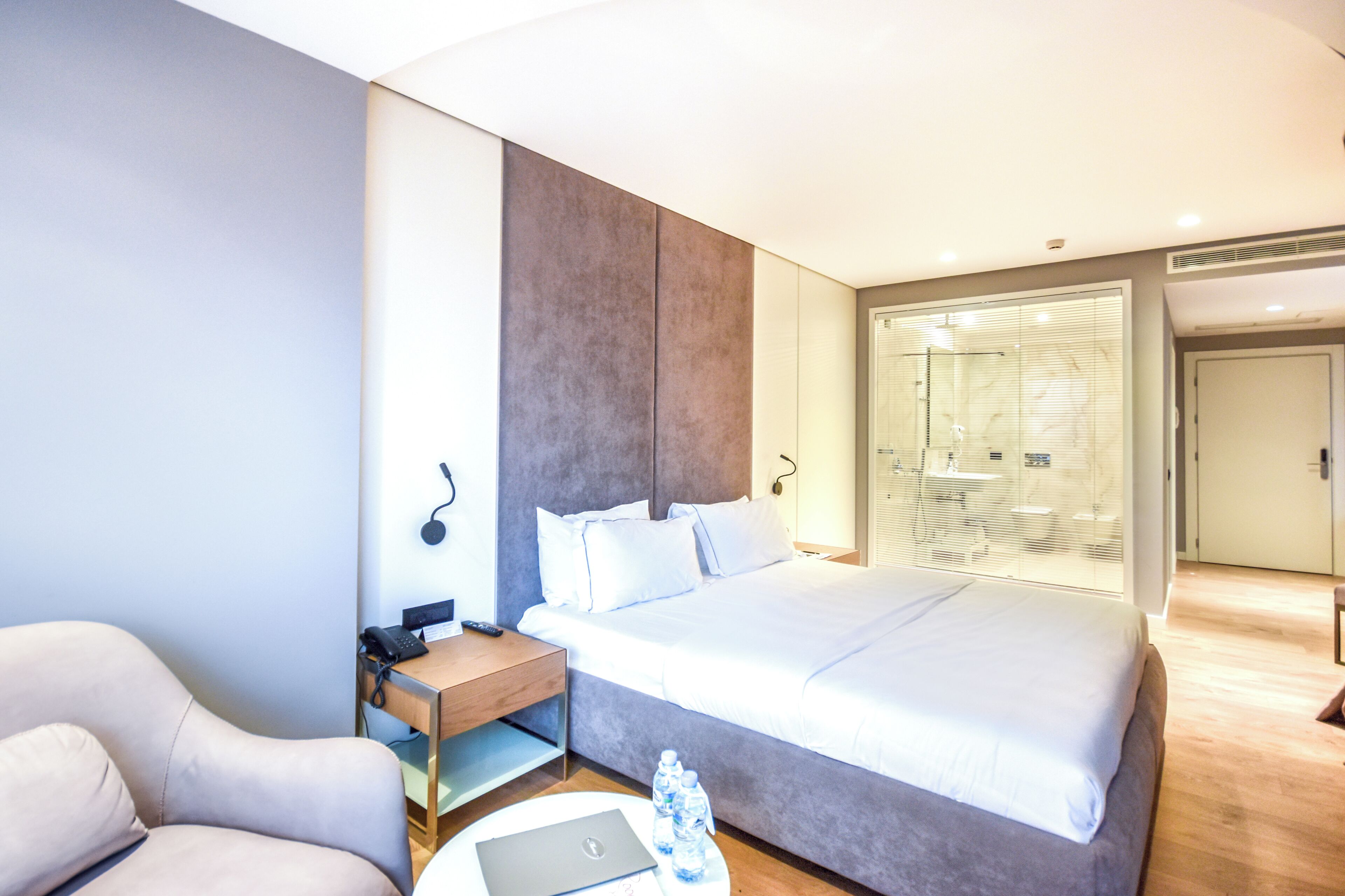 A modern hotel room with a large bed, a desk with a laptop, a chair, and a glass shower area.