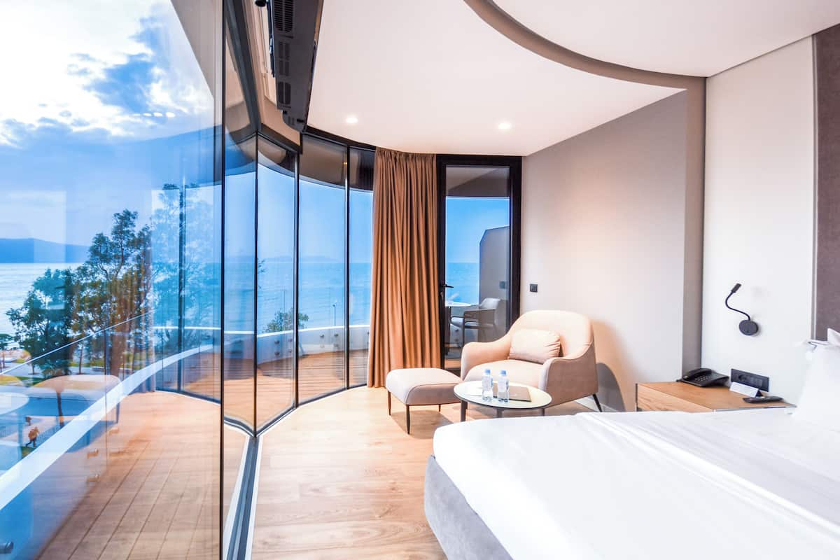 A modern hotel room with a large bed, a seating area with a chair and a small table, a desk with a computer, and a view of the sea through large windows.