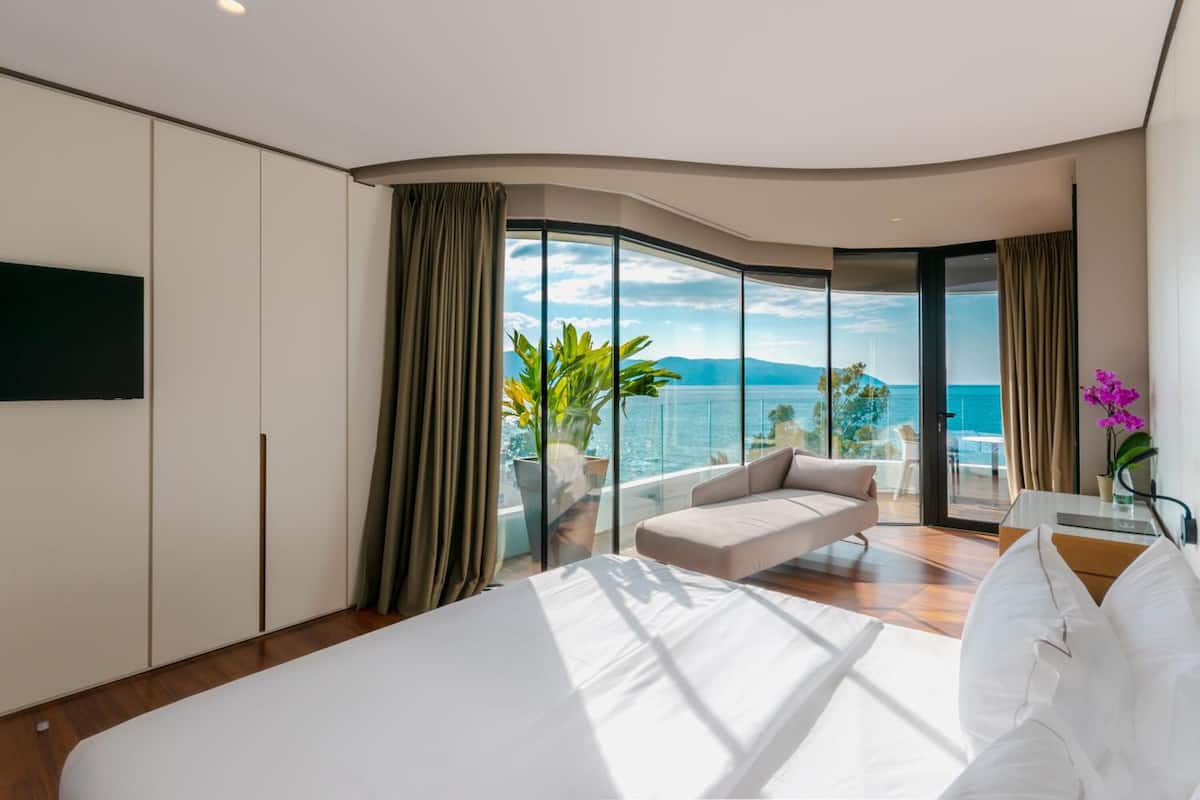 A modern hotel room with a large bed, a sofa, a TV, and a balcony with a view of the sea.