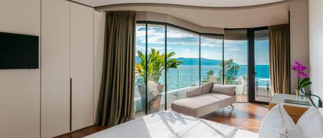 A modern hotel room with a large bed, a sofa, a TV, and a balcony with a view of the sea.