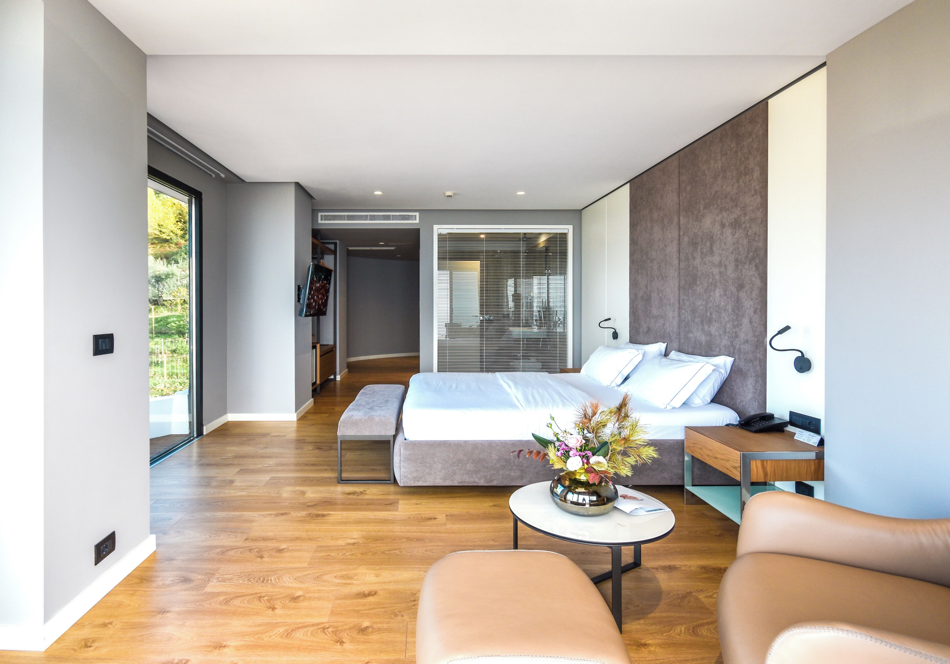 A modern bedroom with a large bed, a bedside table, a small round table with a vase, and a leather armchair.