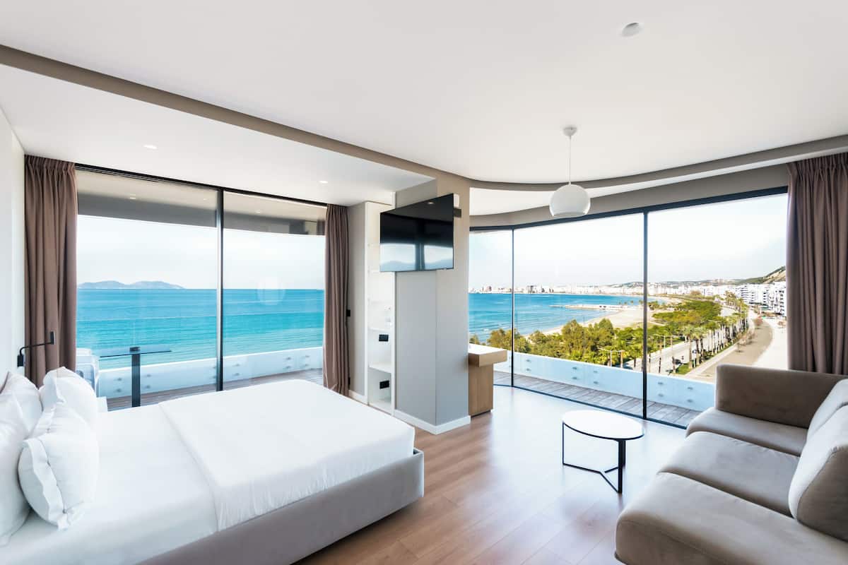 A modern hotel room with a large bed, a sofa, a TV, and a balcony with a sea view.