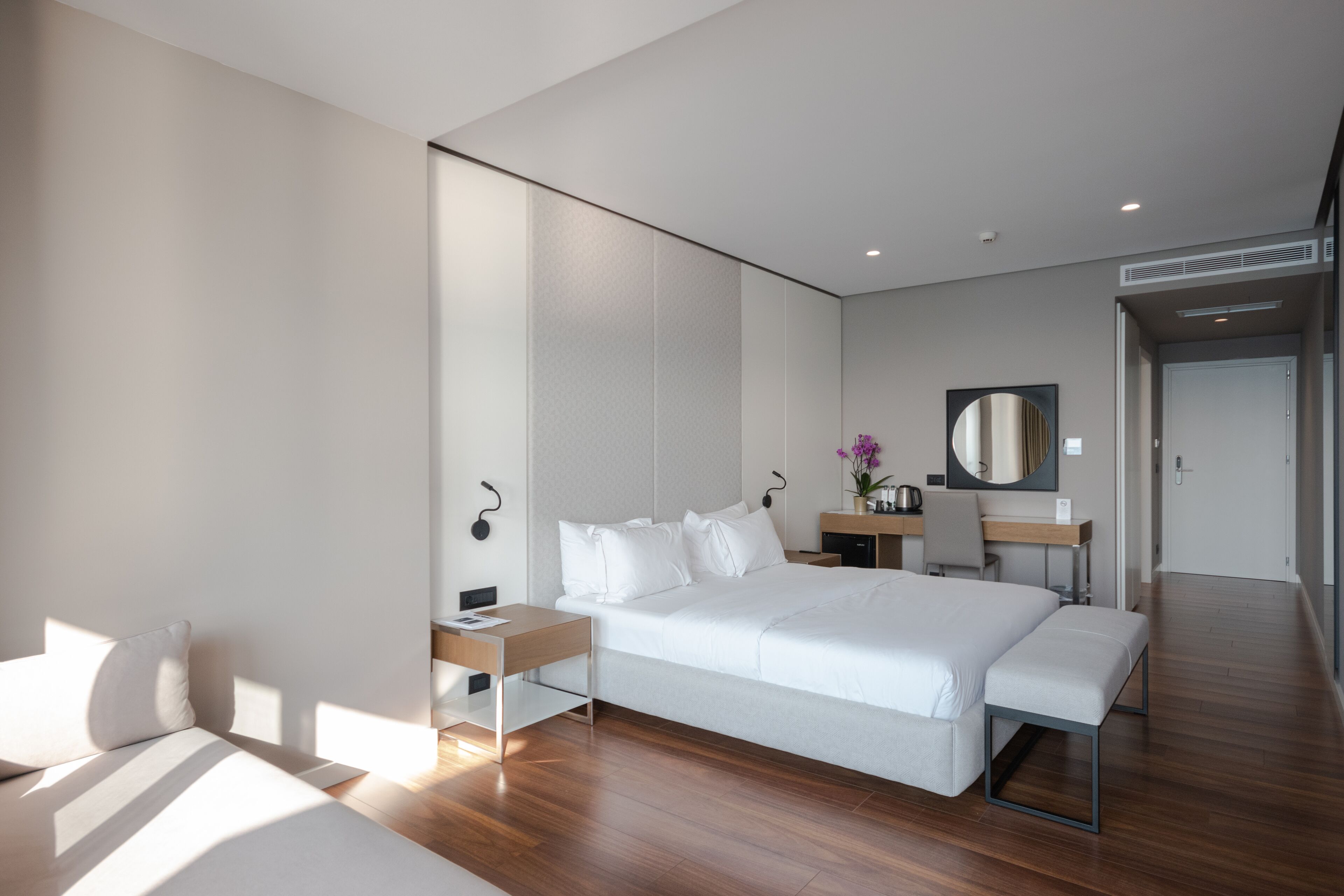 Ein modernes Hotelzimmer mit einem großen Bett, Nachttischen, einem Schreibtisch, einem Sessel, einem Spiegel und einem kleinen Badezimmerbereich.