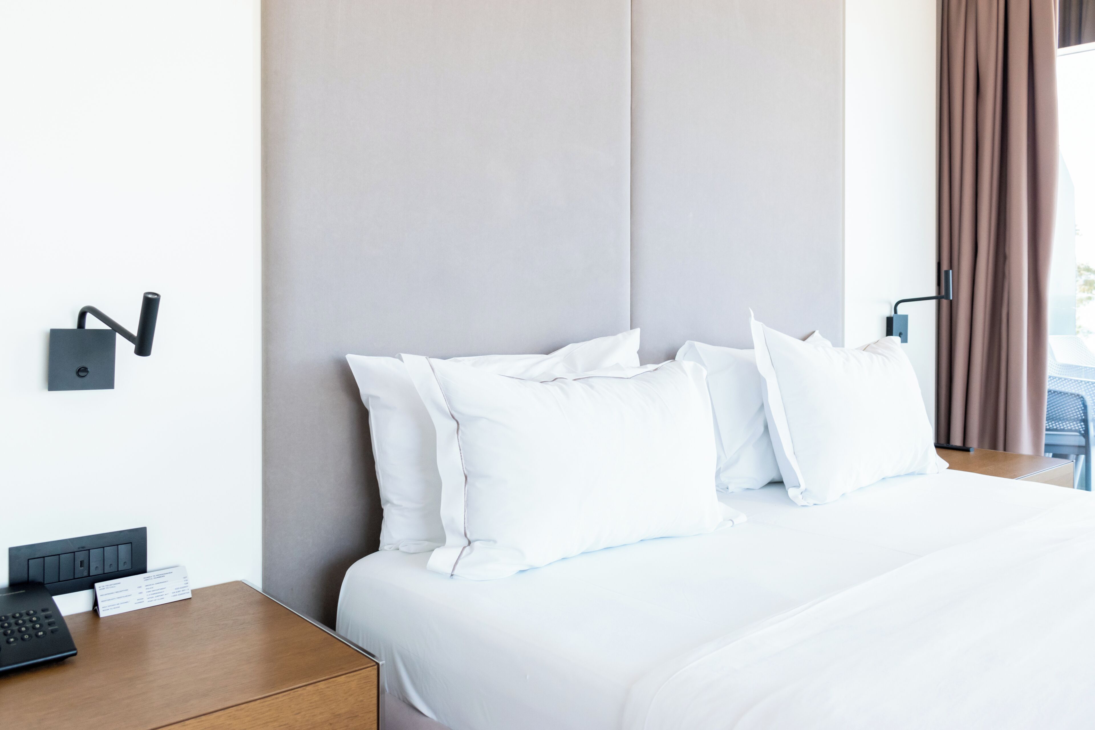 A hotel room with a neatly made bed, a bedside table with a phone and a sign, and a wall-mounted control panel.