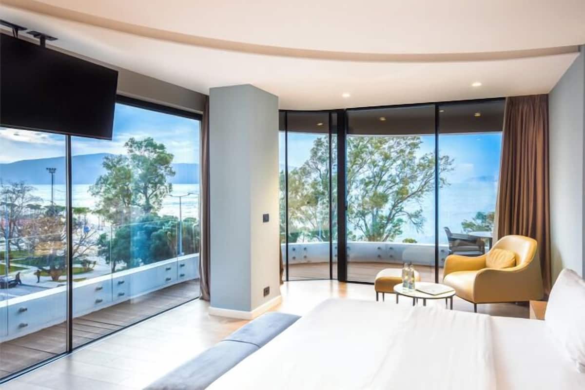 A modern hotel room with a large bed, a sofa, a chair, and a TV. The room has large windows with a view of trees and mountains.