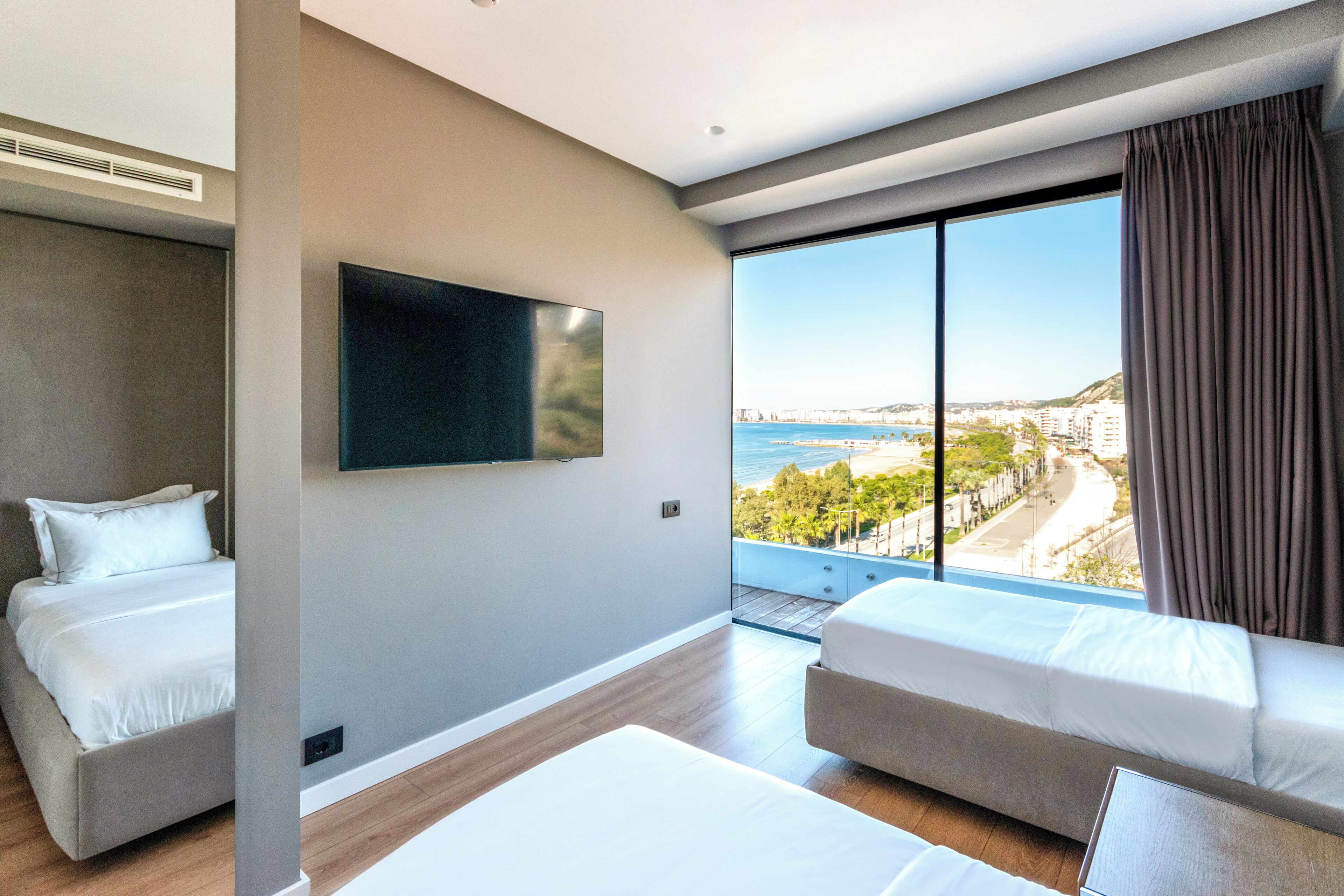 Una habitación de hotel moderna con dos camas, un televisor de pantalla plana y un amplio ventanal con vistas a la ciudad y la playa.