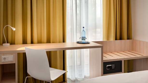 A hotel room with a wooden desk, a white chair, a bedside table with a bottle, and a safe. - Super 8 by Wyndham Augsburg (Augsburg)