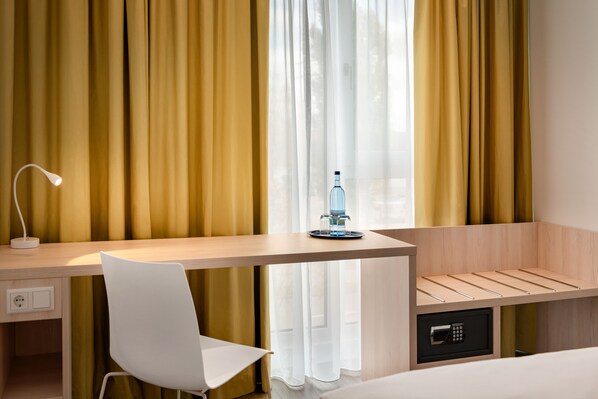 Een hotelkamer met een houten bureau, een witte stoel, een nachtkastje met een fles en een kluis.
