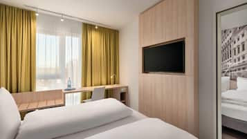 A hotel room with a bed, a desk, a chair, a TV mounted on the wall, and a window with curtains.