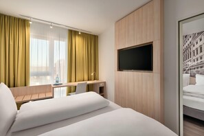 A hotel room with a bed, a desk, a chair, a TV mounted on the wall, and a window with curtains.