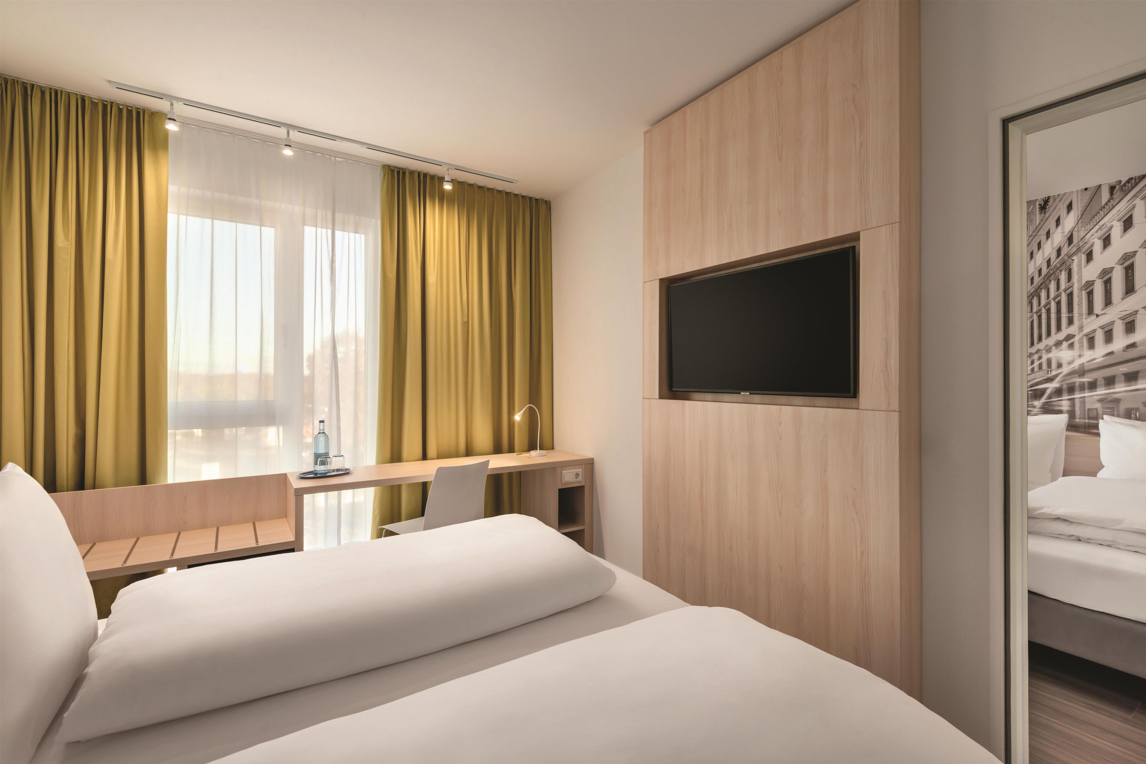 A hotel room with a bed, a desk, a chair, a TV mounted on the wall, and a window with curtains.