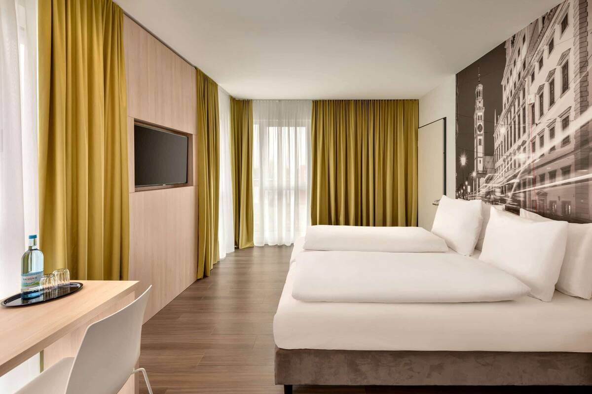 A hotel room with a bed, a desk, a chair, a TV, and a large window with curtains.
