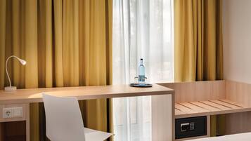 Habitación de hotel con escritorio, silla y cama. Cuenta con una ventana con cortinas, una lámpara y una botella sobre el escritorio.
