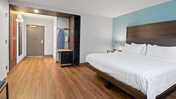 Una habitación de hotel moderna con una cama grande, suelo de madera, una pared de acento azul y una puerta que da a un pasillo con perchero y una silla azul.