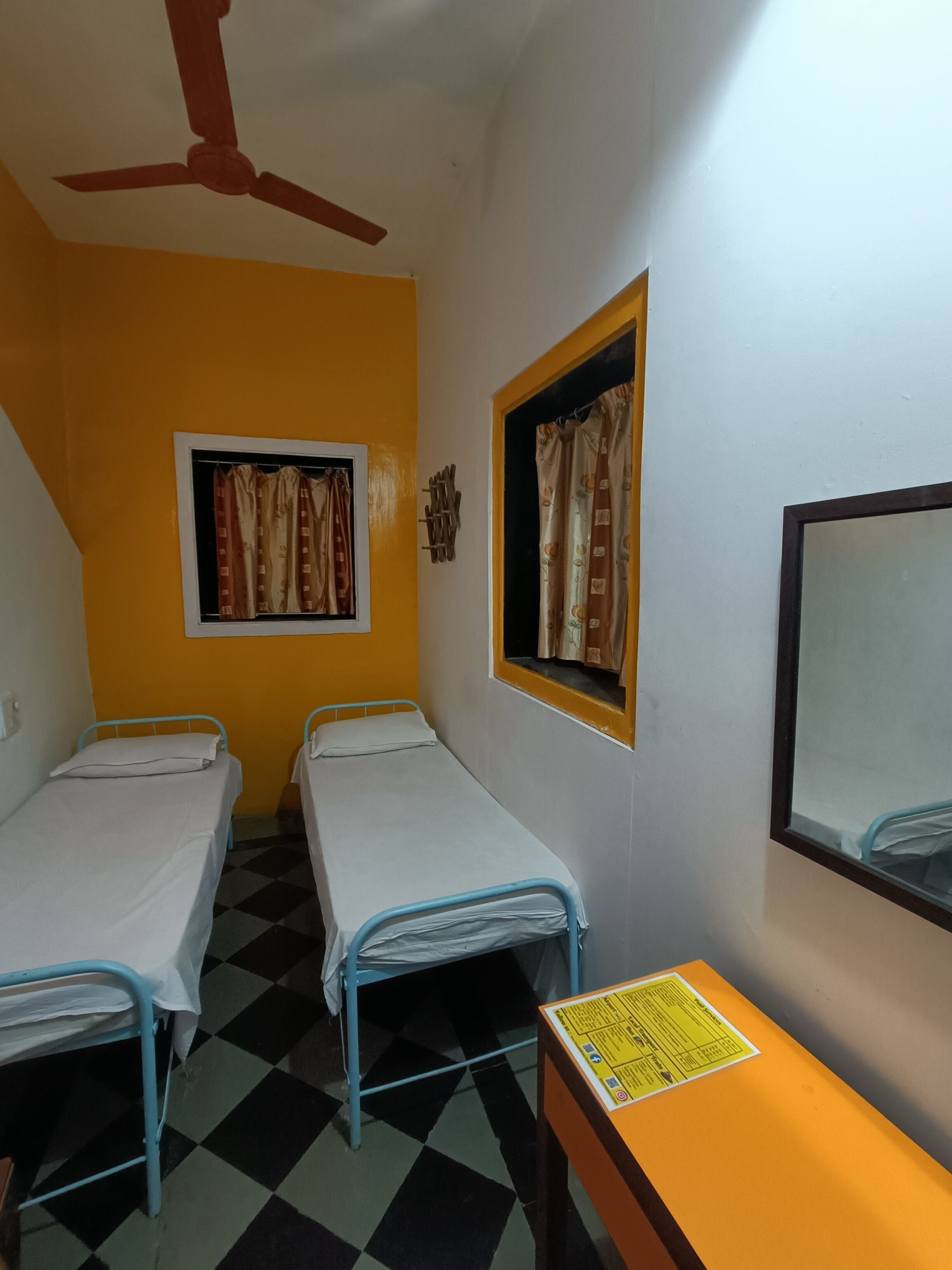 A small, single-room accommodation with two single beds, a ceiling fan, a window with curtains, and a small table with a sign.