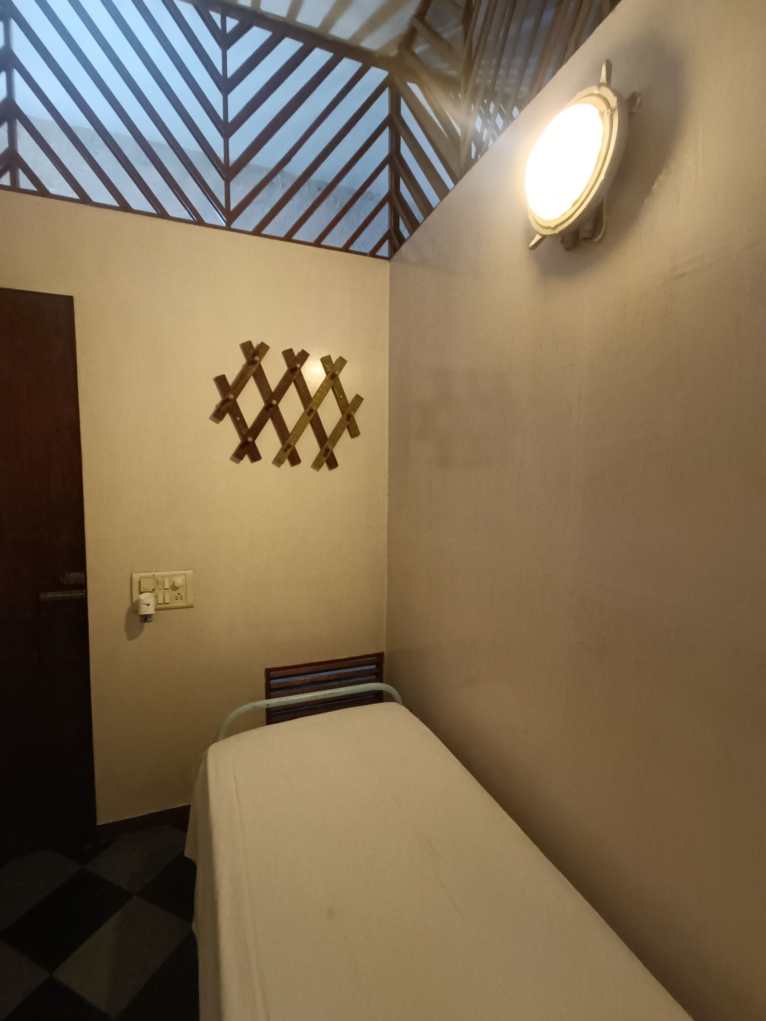 Um quarto pequeno e pouco iluminado, com uma cama de solteiro, uma luminária na parede e um ornamento geométrico na parede.