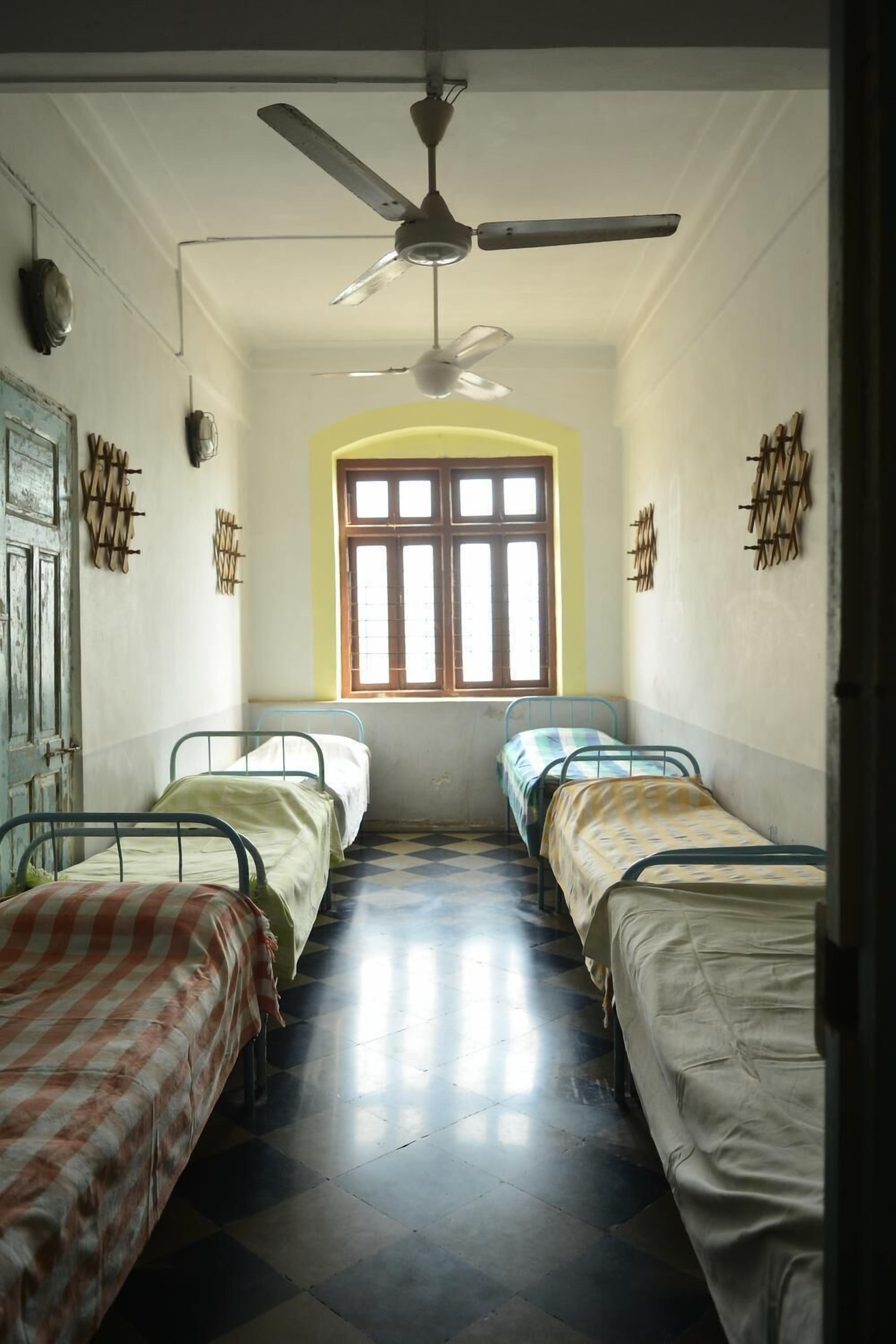A dormitory room with two single beds, a ceiling fan, a window with wooden shutters, and a checkered floor.