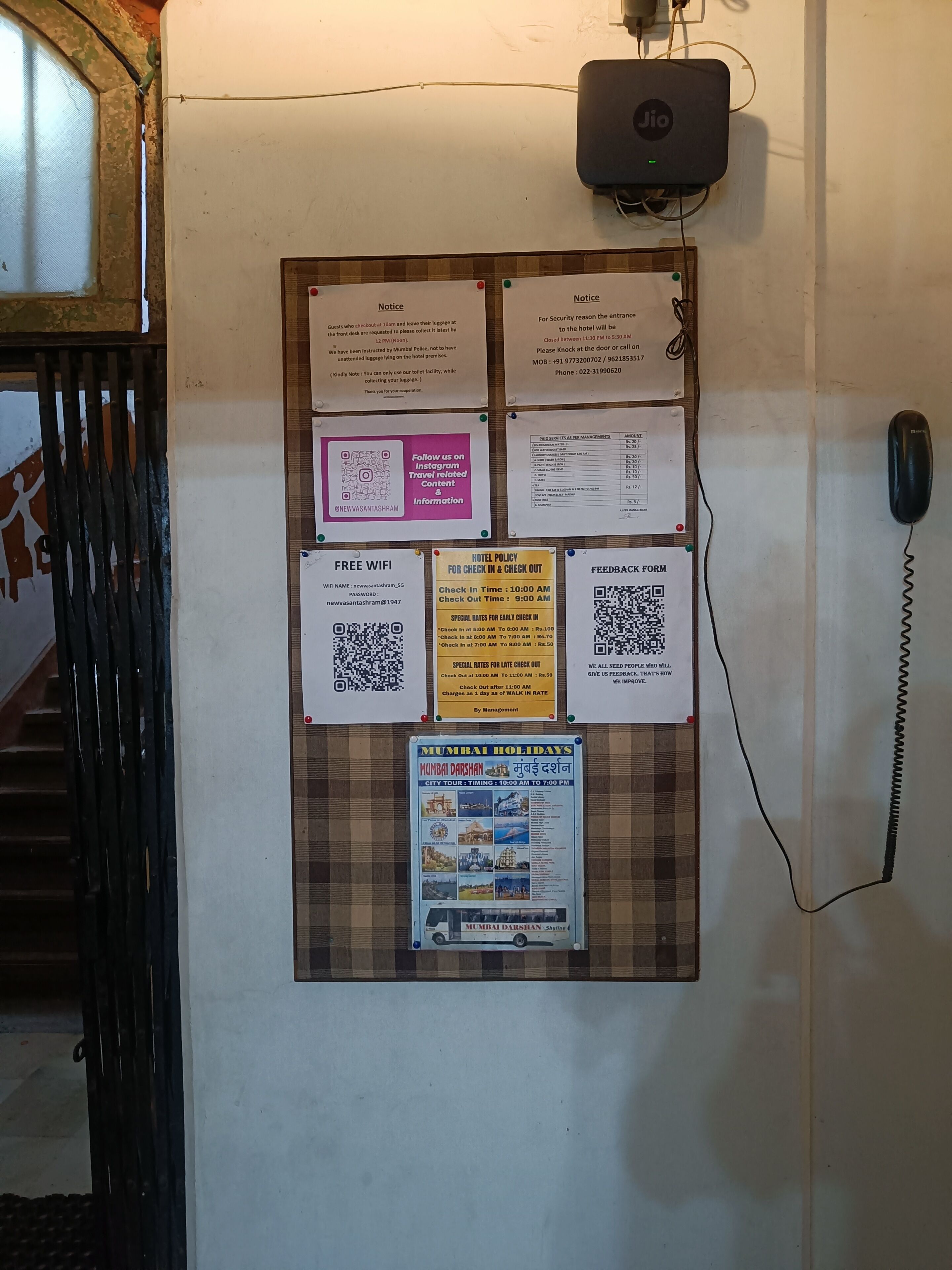 A bulletin board with various notices and information for hotel guests, including check-in and check-out times, WiFi details, and a Mumbai holidays poster.