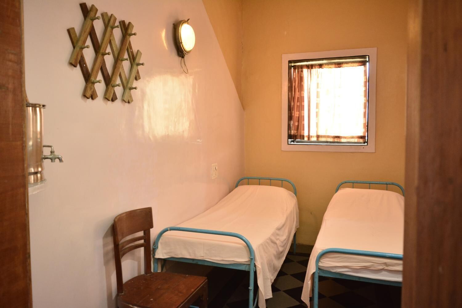 A small, single-room accommodation with two single beds, a wooden chair, a wall-mounted light fixture, and a window with curtains.