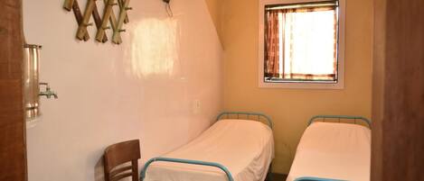 A small, single-room accommodation with two single beds, a wooden chair, a wall-mounted light fixture, and a window with curtains.