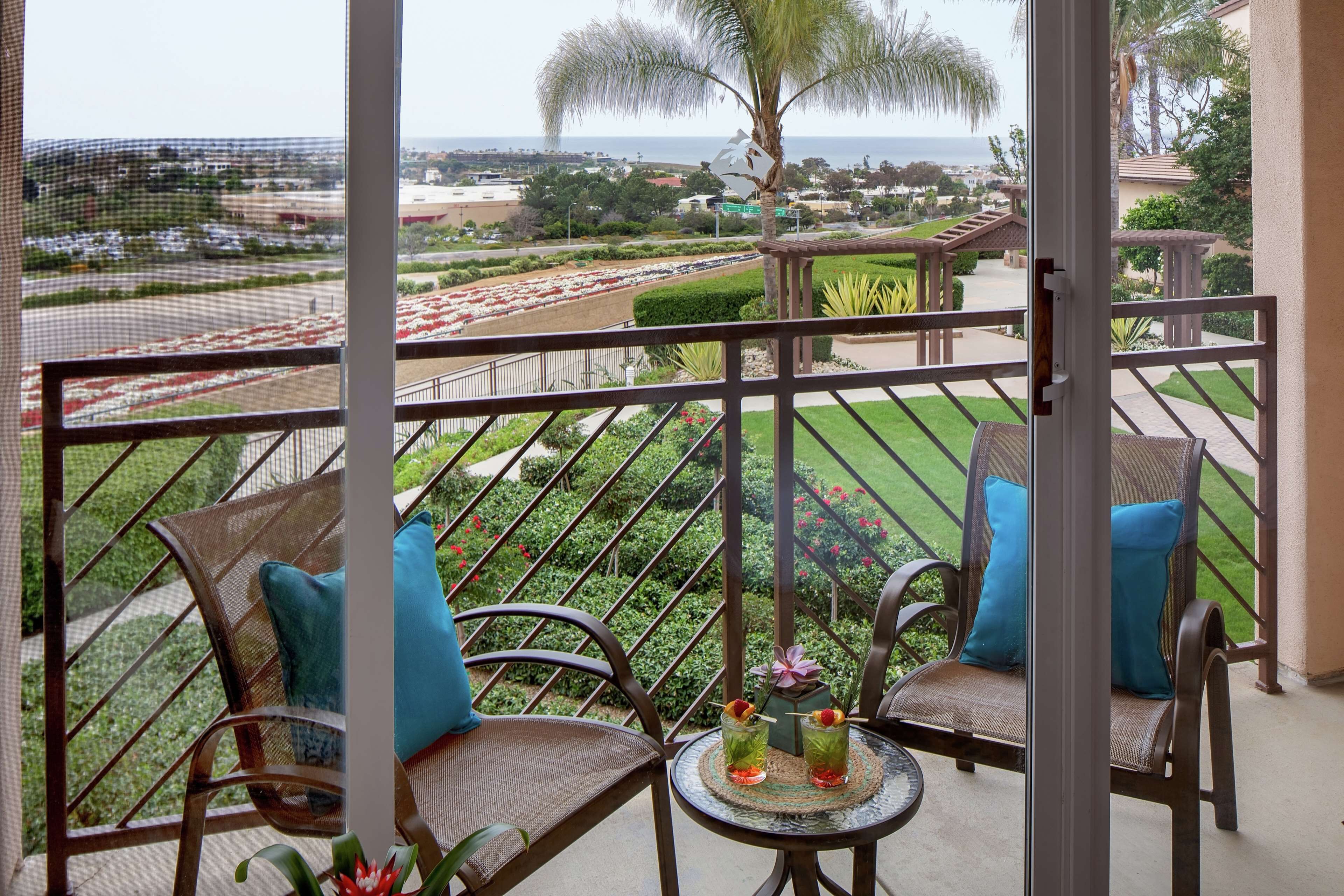 A balcony with wicker chairs and a small table, overlooking a landscaped garden and a road with parked cars.