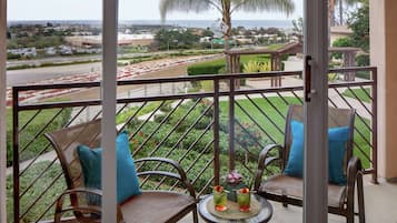A balcony with wicker chairs and a small table, overlooking a landscaped garden and a road with parked cars.