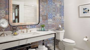 A modern bathroom with a tiled backsplash, a large mirror, a sink with a marble countertop, a toilet, and a framed artwork on the wall.