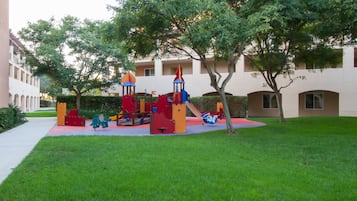 A playground with colorful play structures and a well-maintained lawn in front of a building with arched windows and balconies.
