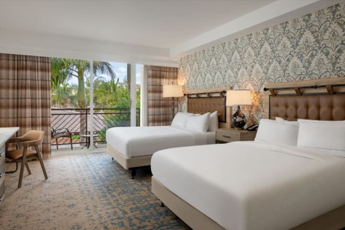 Habitación de hotel con dos camas, un escritorio y una silla. Tiene un balcón con vista a palmeras.
