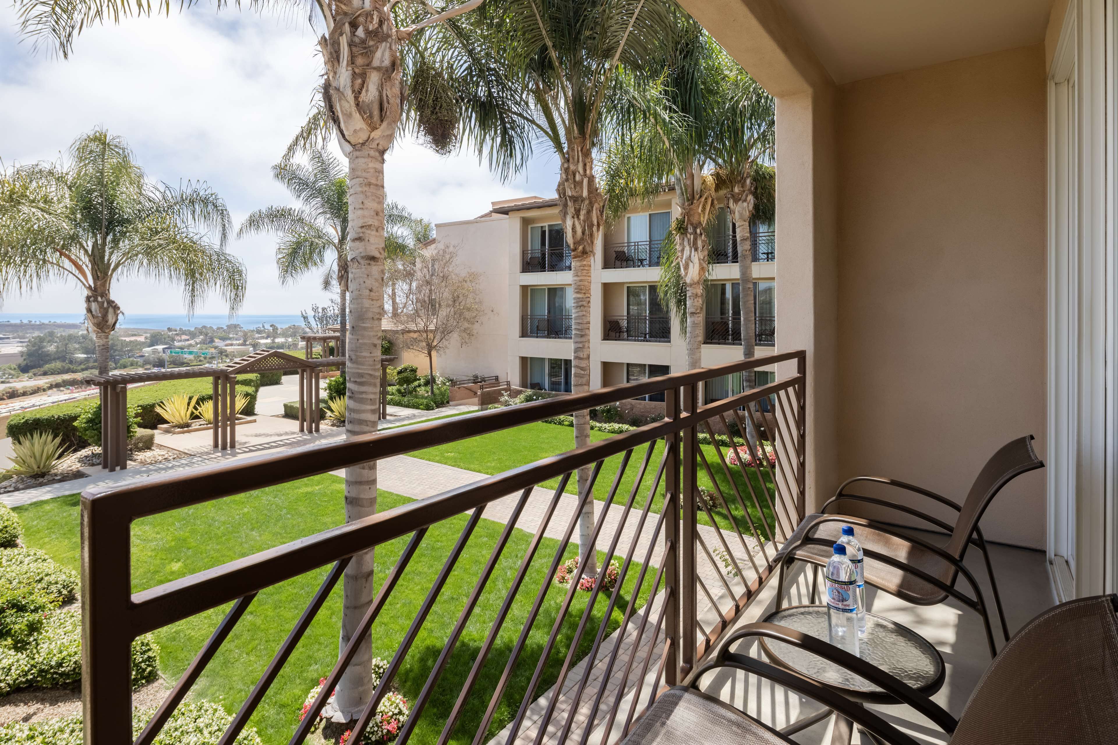 A balcony with a glass table and chairs, overlooking a well-maintained garden and a building with multiple balconies.