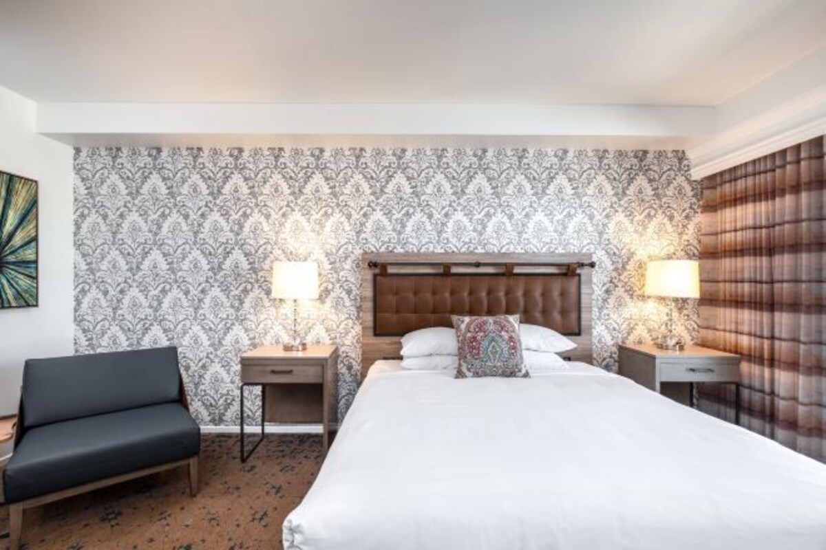 Habitación de hotel con una cama grande, dos mesitas de noche con lámparas, una silla y papel tapiz estampado.
