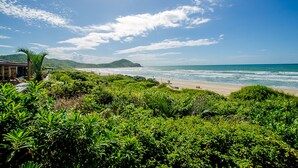 A beach with lush greenery in the foreground, a sandy beach, and a clear blue sky with scattered clouds. - Kalani Pousada (Imbituba)